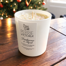 Load image into Gallery viewer, White Perrie Victoria Frankincense &amp; Myrrh candle topped with pale crystals on a wooden table beside a Christmas tree.
